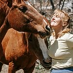 A joyful woman interacts with a horse in a sunny outdoor setting.