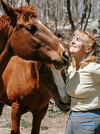 A joyful woman interacts with a horse in a sunny outdoor setting.